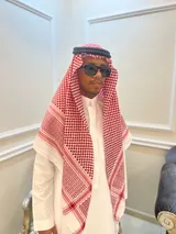 منصور يرتدي ثوبا أبيض وشماغا أحمر وأبيض وعقالا أسود ونظارة سوداء، ويقف في غرفة أنيقة.